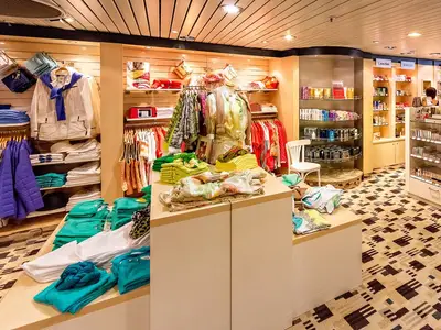 Innenansicht eines gut beleuchteten Shops an Bord der MS Artania. Regale voller Kleidung, Parfüm und Accessoires sind zu sehen. Eine Schaufensterpuppe trägt ein sommerliches Outfit, während rechts ein Bereich für Düfte mit Marken wie Calvin Klein und Gucci erkennbar ist. Der Boden hat ein modernes Muster.