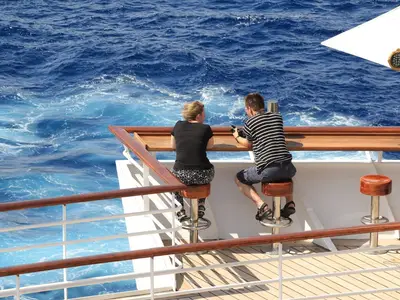 Eine Frau und ein Mann sitzen an einer Holztheke auf dem Sonnendeck der MS Artania und blicken auf das weite, blaue Meer. Die Frau trägt Schwarz, der Mann ein gestreiftes T-Shirt. Ein sonniger Tag an Bord.