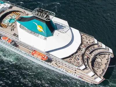 Luftaufnahme der MS Artania, die auf offener See fährt. Zu sehen sind die oberen Sonnendecks mit zwei Pools, Rettungsbooten und zahlreichen Passagieren, die in der Sonne entspannen.