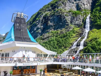Passagiere auf dem Sonnendeck der MS Artania blicken auf einen großen Wasserfall, der von grünen Bergen in einem Fjord herabfliesst. Der Schornstein des Schiffes ist links im Bild.