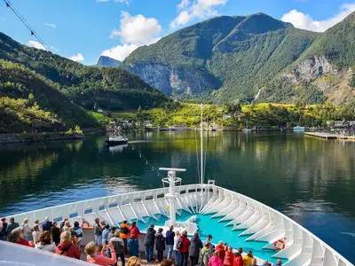 Blick vom Bug der MS Artania: Passagiere genießen die Fahrt durch einen norwegischen Fjord. Grüne Berge, ein Dorf und ein weiteres Schiff sind sichtbar.