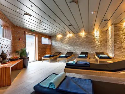 Ein heller und moderner SPA-Ruheraum an Bord der MS Artania. Die Wände sind teilweise aus Holz mit runden Aussparungen und teilweise aus hellem Naturstein. Es gibt mehrere Liegen mit blauen Handtüchern. Zwei runde Fenster zeigen einen hellen Himmel. Eine Pflanze und eine Buddha-Statue dekorieren den Raum.