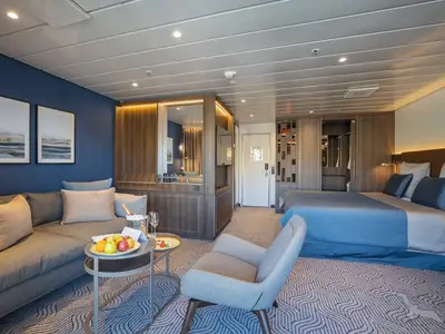 Helle, moderne 2-Bett-Suite auf der MS Artania mit grauem Sofa, Couchtisch mit Obst und Champagner, sowie einem Schlafbereich mit großem Bett und blauer Tagesdecke.