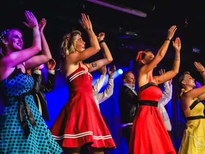Eine Gruppe von Tänzern in farbenfrohen Kleidern und mit Hochsteckfrisuren performt eine lebhafte Show auf der Bühne der MS Artania. Sie strecken die Arme in die Höhe, umgeben von blauer Bühnenbeleuchtung und angedeutetem Publikum. Die Darbietung wirkt energiegeladen und unterhaltsam.