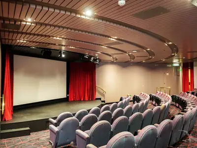 Ein Kinosaal an Bord der MS Artania mit einer großen weißen Leinwand, roten Vorhängen, einer Bühne und leeren Reihen gepolsterter Sitze. Die moderne Decke ist beleuchtet.