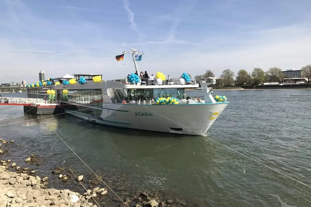 Die MS Asara liegt festlich mit blauen und gelben Ballons geschmückt an einem Flussanleger. Gruppen von Menschen sind auf dem Sonnendeck zu sehen. Im Hintergrund Bäume, Wasser und eine Stadtkulisse.