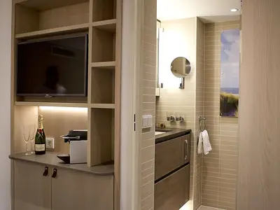 Blick auf das moderne Badezimmer mit Waschtisch und einen Minibar-Bereich mit TV, Kaffeemaschine und Champagnerflasche an Bord der MS Asara. Beige Fliesen, Holzmöbel.