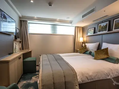 Helle Außenkabine an Bord der MS Asara mit Doppelbett, weißer Bettwäsche und dekorativen Kissen. Links ein Schreibtisch mit TV und einem Fenster mit Jalousien, das Tageslicht hereinlässt.