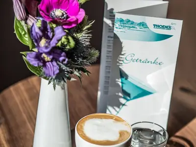 Auf einem Holztisch an Bord der MS Asara stehen eine Kaffeetasse mit Milchschaum, ein Glas und ein Blumengesteck mit lila Blüten. Im Hintergrund ist eine Getränkekarte von Phoenix Reisen zu sehen.