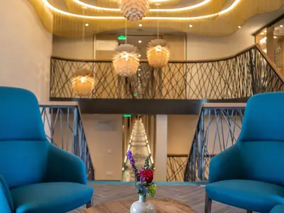 Atrium an Bord der MS Asara mit türkisen Sesseln. Kunstvolles Treppengeländer, Pendelleuchten und ein beleuchteter Gang sind sichtbar. Ein Blumengesteck im Vordergrund.