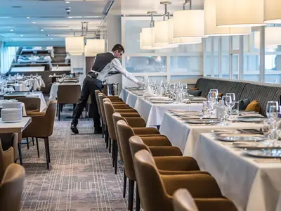 Ein Kellner in schwarzer Weste deckt einen langen Tisch im Restaurant Vier Jahreszeiten an Bord der MS Asara. Die Tische sind mit weißen Tischdecken, Tellern und Gläsern gedeckt. Braune Stühle stehen bereit.
