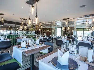 Blick in das elegant gedeckte Restaurant an Bord der MS Asara. Zahlreiche Tische sind bereit mit weißen Tischdecken, Geschirr und Gläsern. Die moderne Inneneinrichtung umfasst viele Hängelampen und eine offene Küche mit grünen Fliesen. Im Hintergrund sind große Fenster mit Blick auf das Wasser zu sehen. Das Restaurant ist menschenleer.