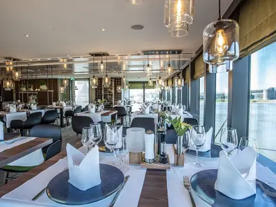 An Bord der MS Asara: Elegantes Restaurant mit vielen gedeckten Tischen und Gläsern. Große Fenster mit Blick auf den Fluss und eine Brücke. Moderne Pendelleuchten.
