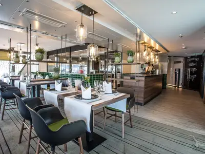 Elegantes Spezialitätenrestaurant der MS Asara. Moderne Tische mit weißer Tischwäsche, schwarze Stühle und grüne Akzente. Die Theke im Hintergrund hat dunkelgrüne Fliesen.
