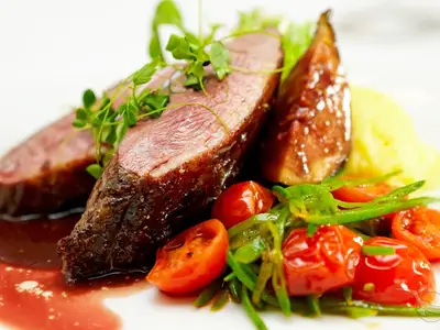Close-up of a perfectly cooked meat dish, possibly duck or beef, garnished with fresh herbs, cherry tomatoes, and green beans, served with a rich red sauce and a side of mashed potatoes, on a white plate aboard the MS Asara.