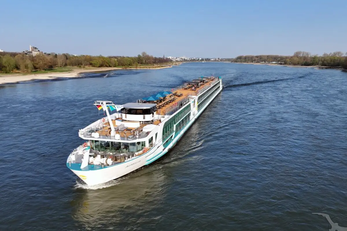 Die MS Aurelia, ein weißes Flusskreuzfahrtschiff, fährt auf einem breiten Fluss. Das Sonnendeck mit Liegen und blauen Schirmen ist sichtbar. Bewaldete Ufer und ferne Gebäude unter blauem Himmel.