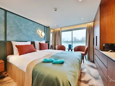 Eine moderne und helle Balkonkabine an Bord der MS Aurelia. Sie bietet ein Doppelbett, zwei Sessel am Fenster mit Flussblick und eine stilvolle Einrichtung in Blau- und Holztönen.