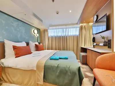 Helle Außenkabine der MS Aurelia. Doppelbett mit weißen und rostfarbenen Kissen, Fenster, Schreibtisch mit TV und Sessel. Moderne Einrichtung mit Holzakzenten und mintfarbenen Wänden.