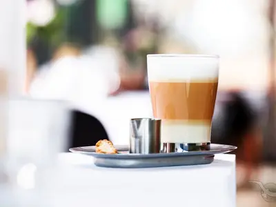 An Bord der MS Aurelia: Eine mehrschichtige Kaffeespezialität in hohem Glas, mit Milchkännchen und Keks auf Tablett. Unscharfer, heller Hintergrund.