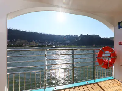 Ein Blick vom Deck der MS Aurelia auf einen breiten Fluss. Im Vordergrund ein glänzendes Schiffsreling und ein orangefarbener Rettungsring. Im Hintergrund ist ein malerisches Dorf mit Häusern und einer Burg auf einem bewaldeten Hügel unter klarem Himmel und strahlender Sonne zu sehen.