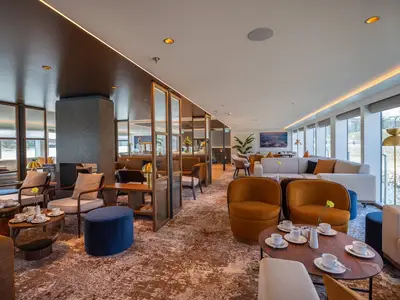 Eine stilvolle, helle Lounge an Bord der MS Aurelia mit modernen Sitzgelegenheiten, Kaffeetischen und großen Panoramafenstern mit Ausblick auf das Ufer.