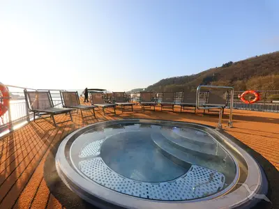 Sonnendeck der MS Aurelia mit einem ovalen Whirlpool im Vordergrund, umgeben von braunen Liegestühlen. Im Hintergrund sind bewaldete Hügel unter blauem Himmel zu sehen.