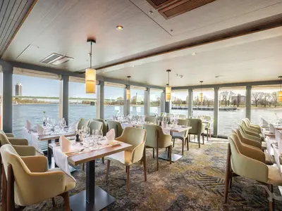 Helles, elegantes Restaurant der MS Aurelia mit weiß gedeckten Tischen und hellgrünen Stühlen. Große Fenster bieten Panoramablick auf einen Fluss, eine Brücke und die Uferlandschaft am Tag. Ohne Personen.