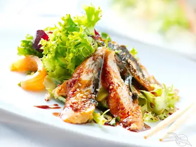 Nahaufnahme eines appetitlichen Salats mit glasierten Aalscheiben, frischem Grünsalat, Mandarinenstücken und Sesam auf weißem Teller an Bord der MS Aurelia, garniert mit dunkler Sauce.