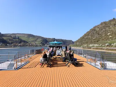 Gäste entspannen auf dem sonnigen Sonnendeck der MS Aurelia, das Schiff gleitet auf einem Fluss vorbei an bewaldeten Hügeln und einem Schloss. Strahlend blauer Himmel.