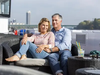 Ein lächelndes Paar mittleren Alters entspannt auf einem bequemen Lounge-Sofa an Bord der MS Aurelia. Im Hintergrund ist eine Stadtsilhouette zu sehen, während die Sonne scheint. Ein erfrischendes Getränk steht bereit.
