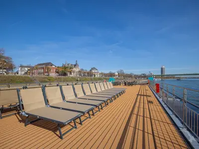 Blick vom Sonnendeck der MS Aurelia mit Liegestühlen. Im Hintergrund sind ein Fluss, eine Stadt und eine Brücke bei strahlendem Sonnenschein zu sehen.
