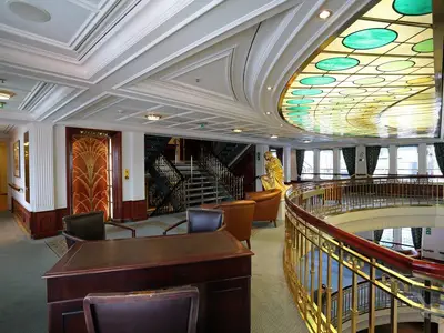 Das opulente Atrium an Bord der MS Deutschland. Eine prächtige Buntglasdecke, eine goldene Statue und ein verzierter Art-Déco-Aufzug prägen den luxuriösen Innenbereich mit Grand Staircase.
