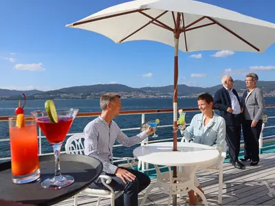 Zwei Paare sitzen und stehen auf dem Sonnendeck der MS Deutschland, trinken Cocktails mit Blick auf das Meer und eine grüne Küstenlandschaft. Ein Sonnenschirm spendet Schatten.