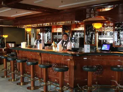 Die Bar 'Zum Alten Fritz' an Bord der MS Deutschland zeigt zwei lächelnde Barkeeper hinter einer holzvertäfelten Theke und Barhockern. Gläser und Flaschen in den Regalen. Klassische, elegante Einrichtung.