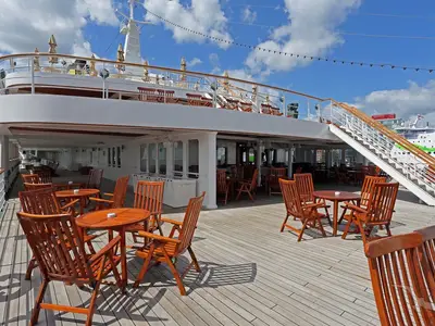 Das Sonnendeck der MS Deutschland mit zahlreichen leeren Holztischen und Stühlen. Eine Treppe führt zum oberen Deck. Im Hintergrund ist ein weiteres Schiff zu sehen.