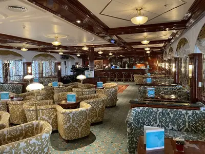 Eine elegante Bar-Lounge an Bord der MS Deutschland mit zahlreichen gemusterten Sesseln und Sofas. Im Hintergrund befindet sich eine holzgetäfelte Bar mit Barhockern und Fenstern.