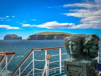 Von Bord der MS Deutschland, eine Bronzeskulptur im Vordergrund. Im Hintergrund das weite Meer mit grünen, felsigen Inseln unter strahlend blauem Himmel.