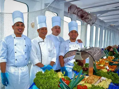 Vier lächelnde Köche in weißen Uniformen und hohen Kochmützen stehen an einem reichhaltigen Buffet mit frischem Salat und Speisen an Bord der MS Deutschland.