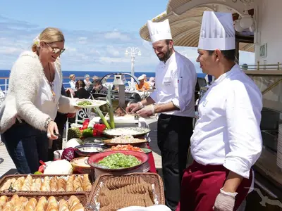 An Bord der MS Deutschland genießen Gäste ein reichhaltiges Buffet an Deck. Eine Frau nimmt sich Essen, während zwei Köche Speisen vorbereiten. Im Hintergrund sind das Meer und weitere Passagiere zu sehen.