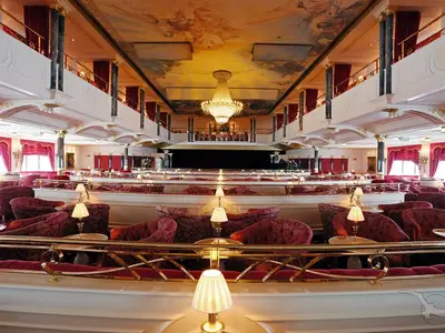 Der prunkvolle Kaisersaal der MS Deutschland mit Bühne, eleganten roten Sesseln, goldenen Balustraden, Deckenmalerei und imposantem Kronleuchter.
