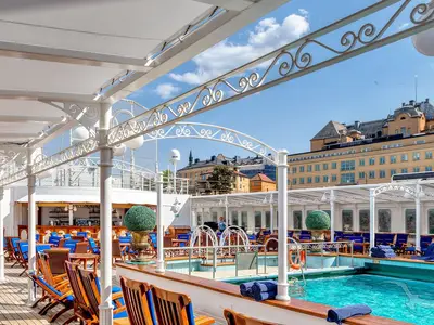 An Bord der MS Deutschland: Das sonnige Pooldeck mit blauem Pool, Liegestühlen und einer Bar. Elegante weiße Pergolen rahmen den Blick auf eine Stadt. Ein Crew-Mitglied ist sichtbar.