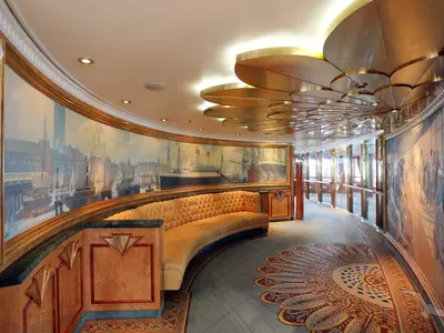 Elegante Promenade an Bord der MS Deutschland. Eine Wandmalerei einer Hafenstadt. Geschwungene Chaiselongue und kunstvolle goldene Deckenverzierung. Der Boden ist mit einem Teppich gemustert.