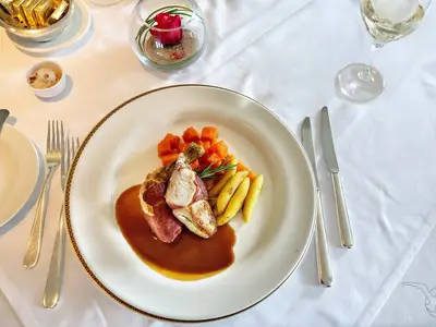 An Bord der MS Deutschland: Elegantes Hauptgericht mit gebratenem Fleisch in Prosciutto, dunkler Sauce und Gemüse auf weißem Teller. Besteck liegt daneben.