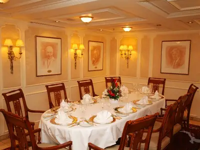 Der Kanzler Zimmer an Bord der MS Deutschland. Ein festlich gedeckter ovaler Tisch mit goldenen Platztellern, weißen Servietten und einem Blumenarrangement. Gerahmte Porträts und goldene Wandleuchten zieren die Wände.