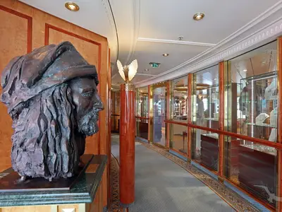 Blick auf einen eleganten Flur an Bord der MS Deutschland mit einer prominenten Bronzebüste im Vordergrund und beleuchteten Boutiquen mit Schmuck und Waren in Vitrinen entlang der Wand.