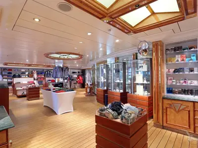 Ein weitläufiger Bord-Shop an Bord der MS Deutschland mit Holzfußboden und hellen Decken. Auslagen präsentieren Kleidung, Schmuck in Vitrinen und Parfüms in Wandregalen. Der Shop ist leer von Personen.