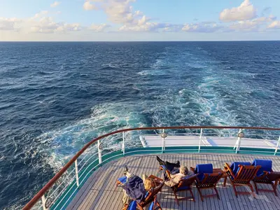 Blick vom Heck der MS Deutschland auf das weite, blaue Meer und die schäumende Heckwelle. Zwei Personen entspannen in Liegestühlen auf dem Holzdeck.