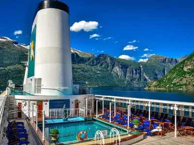 Das Sonnendeck der MS Deutschland zeigt einen Pool und blaue Liegestühle. Im Hintergrund erheben sich grüne Berge mit schneebedeckten Gipfeln über einem tiefblauen Fjord unter klarem Himmel.