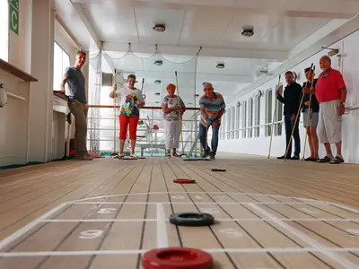 Acht Personen spielen Shuffleboard auf dem Holzdeck der MS Deutschland. Spielscheiben und Markierungen des Spielfeldes sind im Vordergrund zu sehen.