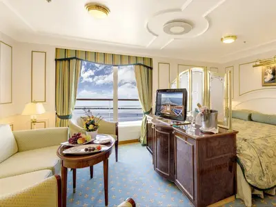 Elegante Suite an Bord der MS Deutschland mit Sofa, Couchtisch mit Obst, großem Fenster mit Meerblick, Fernseher auf einem Schrank und einem Teil des Bettes.